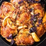 Glazed bone-in, skin-on chicken thighs with caramelized apple wedges, shallots, and pancetta, garnished with fresh thyme and purple basil, overhead close-up in a black cast iron skillet on a dark background.