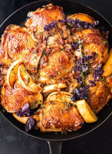 Glazed bone-in, skin-on chicken thighs with caramelized apple wedges, shallots, and pancetta, garnished with fresh thyme and purple basil, overhead close-up in a black cast iron skillet on a dark background.