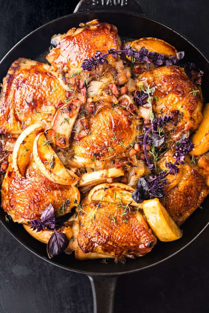Glazed bone-in, skin-on chicken thighs with caramelized apple wedges, shallots, and pancetta, garnished with fresh thyme and purple basil flowers, overhead close-up in a black cast iron skillet on a dark background.