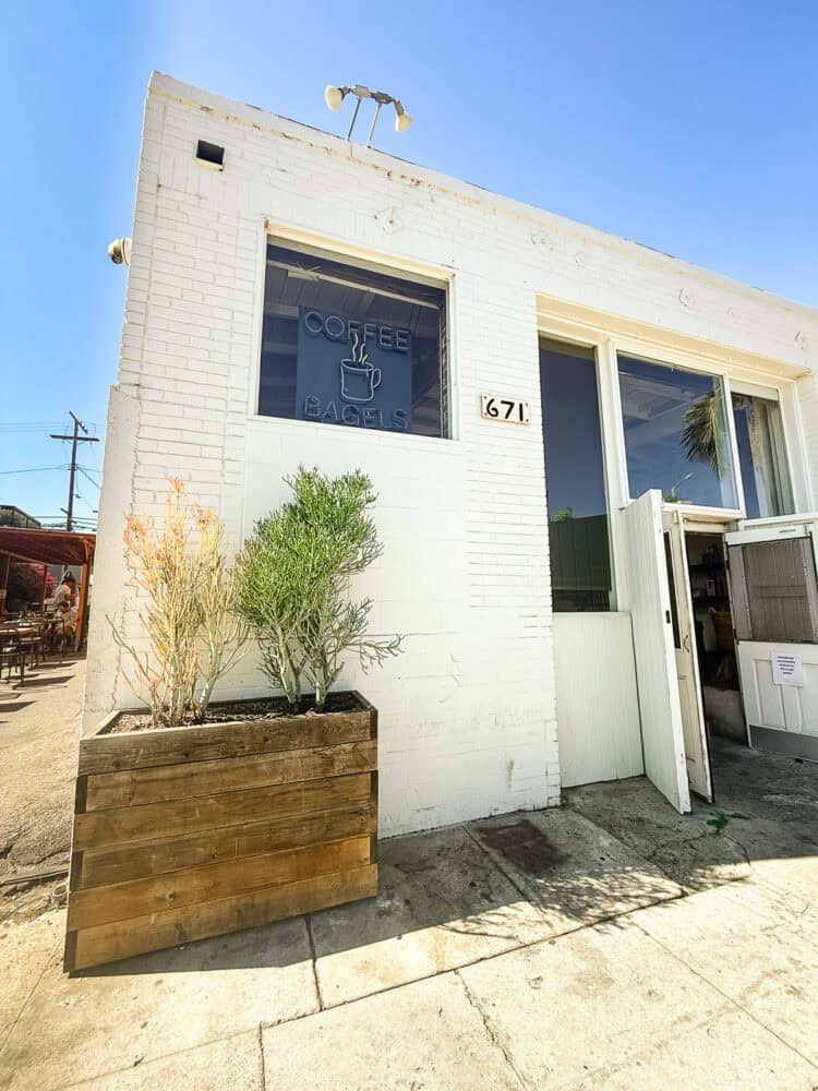 White brick exterior of Groundwork Coffee on Rose Ave in Venice Beach, with a neon "Coffee Bagels" sign in the window and a wooden planter out front.