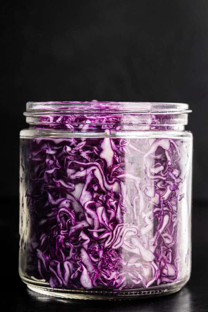 Thinly sliced red cabbage, that actually looks purple, packed into a glass jar before the brine is poured in, on a dark background, close-up angle.