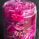 Pickled red cabbage, the brine turned a bright magenta from the vinegar, with visible mustard seeds, black peppercorns, and a bay leaf, packed into a glass jar on a dark background, close-up angle.
