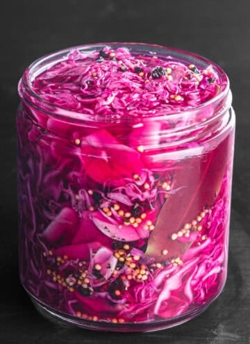 Pickled red cabbage, the brine turned a bright magenta from the vinegar, with visible mustard seeds, black peppercorns, and a bay leaf, packed into a glass jar on a dark background, close-up angle.