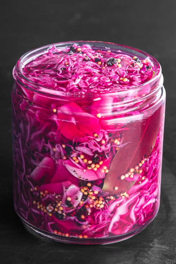 Pickled red cabbage, the brine turned a bright magenta from the vinegar, with visible mustard seeds, black peppercorns, and a bay leaf, packed into a glass jar on a dark background, close-up angle.