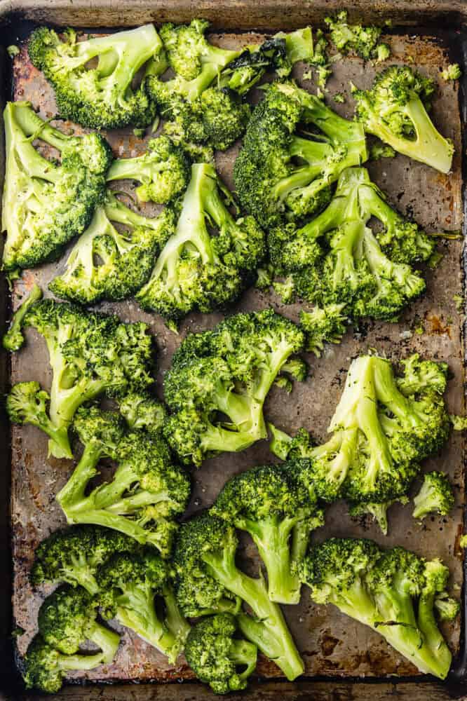 Overhead shot of bright green steamed broccoli florets smashed flat in a single layer on a dark, well-worn baking sheet, ready to be roasted.