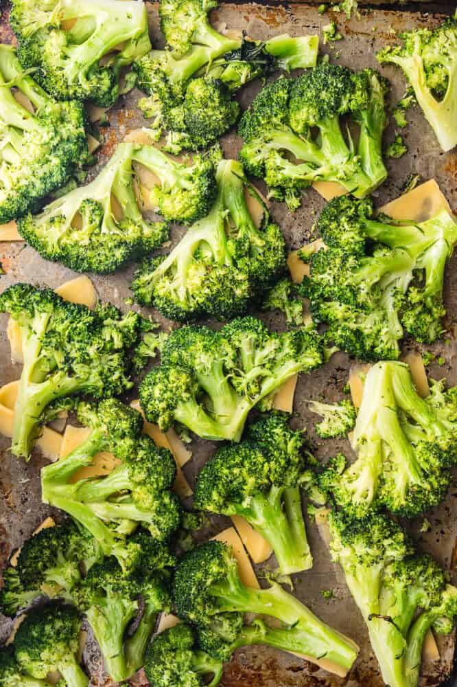 Overhead shot of smashed bright green broccoli florets with slices of cheese tucked under them on a baking sheet before roasting.